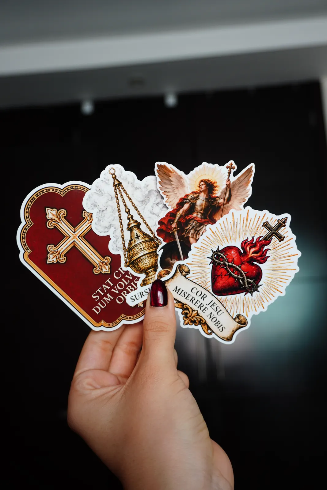 Mano sosteniendo cuatro stickers troquelados: Stat Crux, incensario, San Miguel Arcángel y Cor Jesu miserere nobis